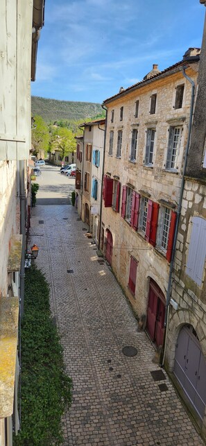 Une ruelle étroite pavée, bordée de bâtiments historiques en pierre aux volets rouges, et un ciel d’un bleu limpide au-dessus.