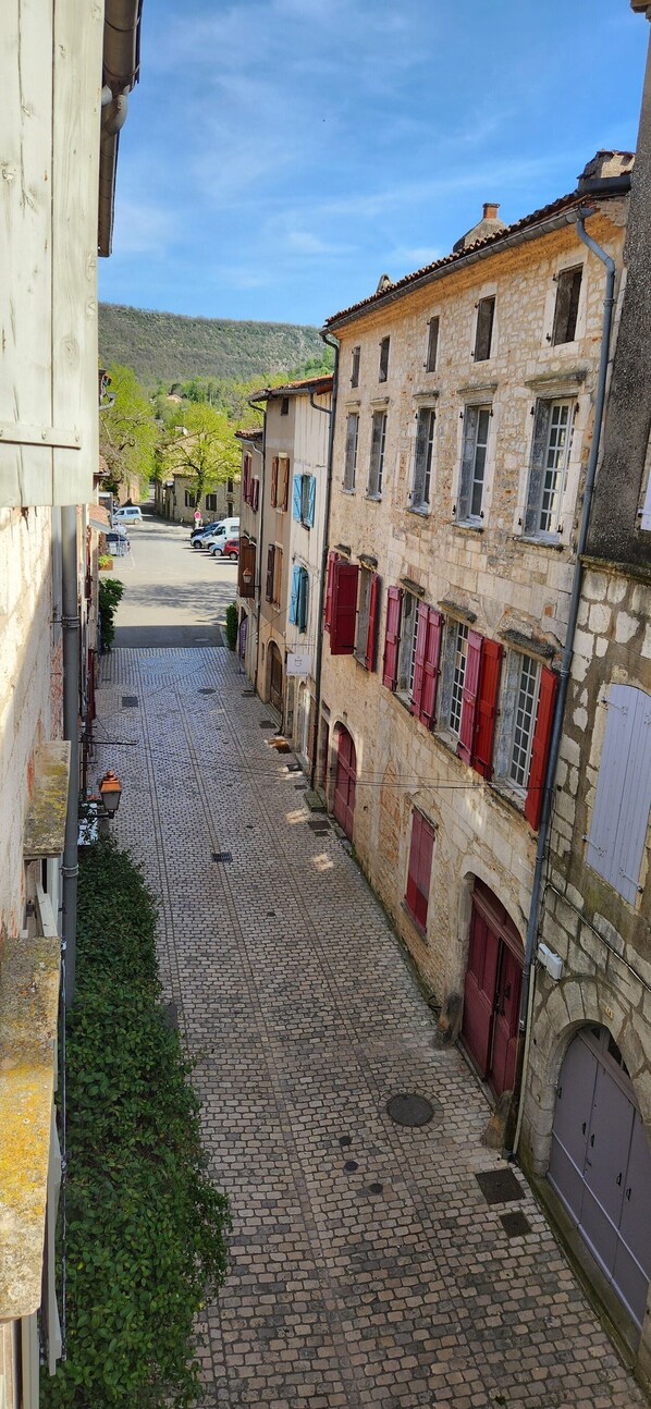 Una estrecha calle empedrada flanqueada por edificios históricos de piedra con contraventanas rojas y un cielo azul despejado.