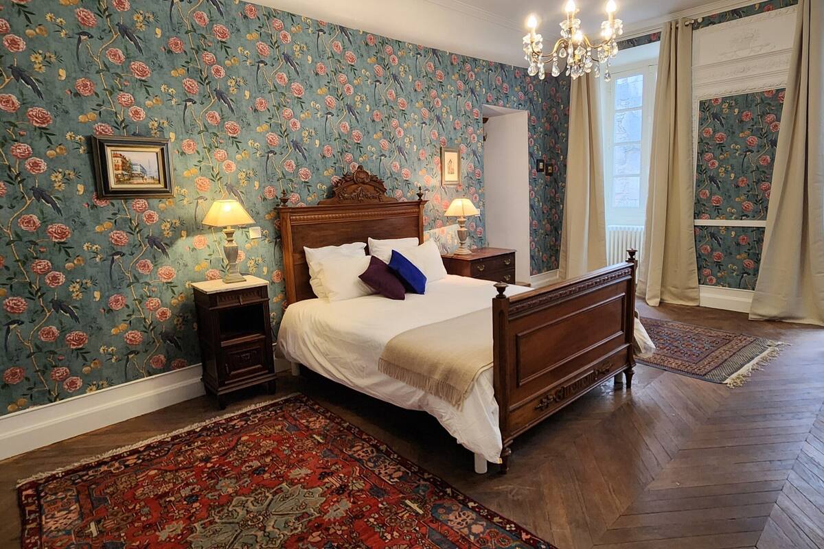Une chambre avec un papier peint à motifs floraux, un lit en bois, deux tables de chevet avec des lampes, un tapis rouge à motifs, et une vue sur l’extérieur par une fenêtre.
