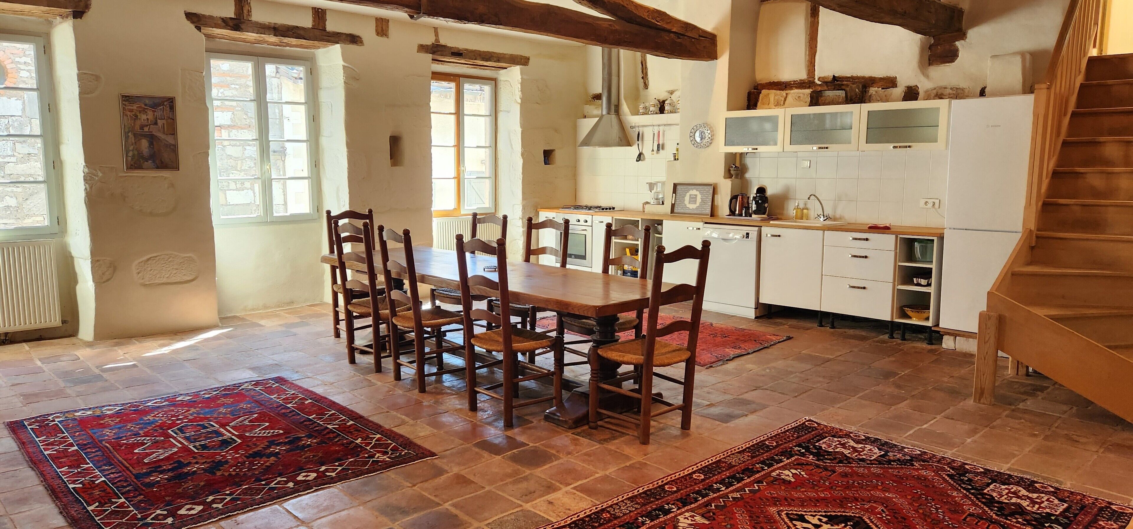 Un grand espace à manger avec une table et des chaises en bois, une cuisine avec des meubles blancs et un escalier.