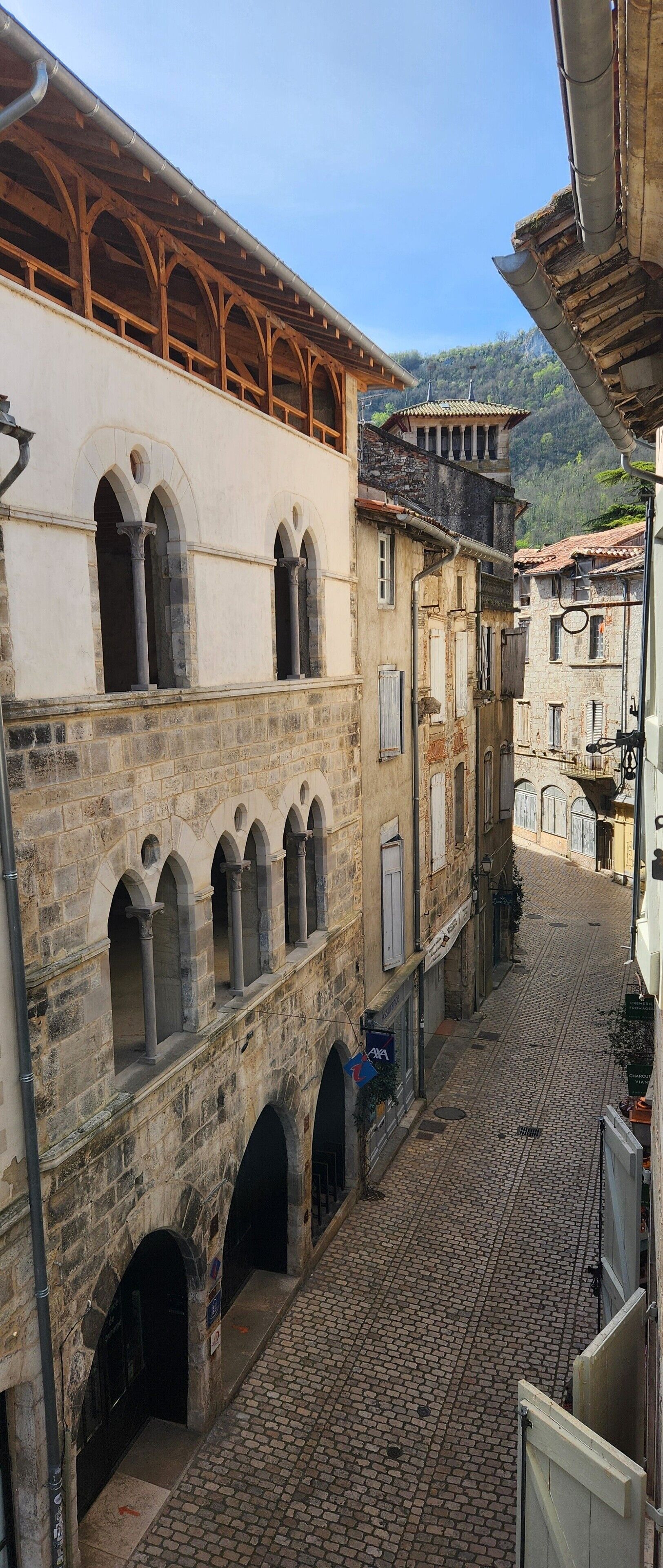 Une ruelle étroite pavée, bordée de bâtiments historiques dotés d’arcs en plein cintre et d’un balcon en bois.