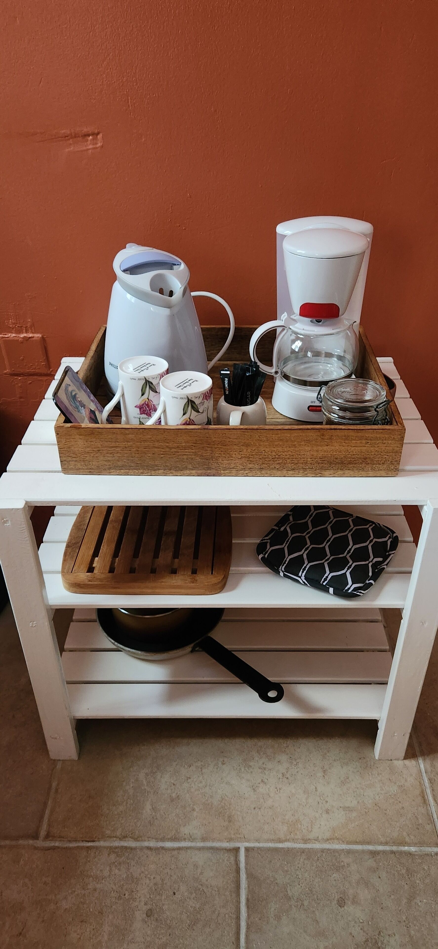 Un coin cuisine comprenant une machine à café, des tasses, une bouilloire, une poêle et une planche à découper, le tout posé sur une étagère blanche.