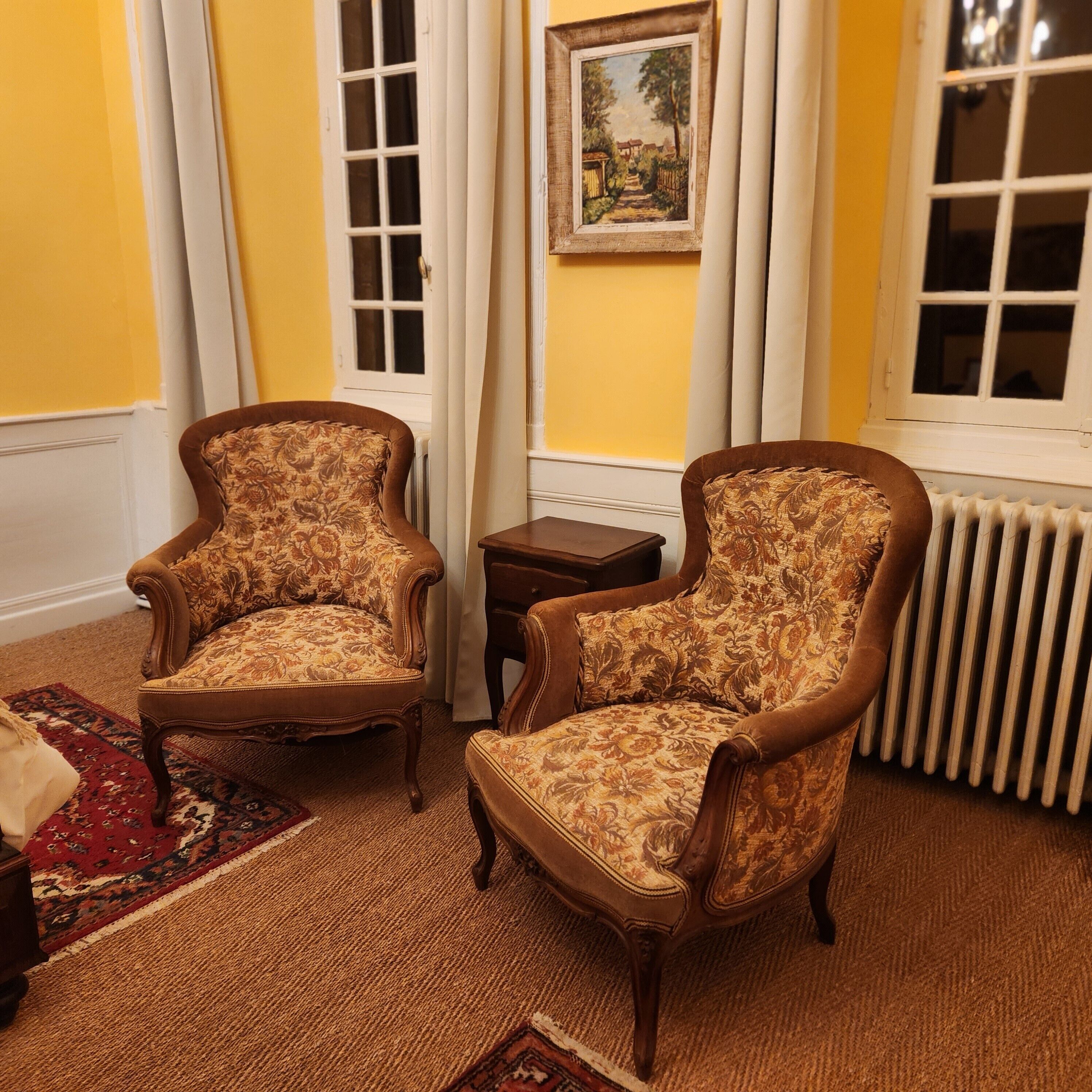A room with two ornate armchairs, a small table, and a painting on the wall.