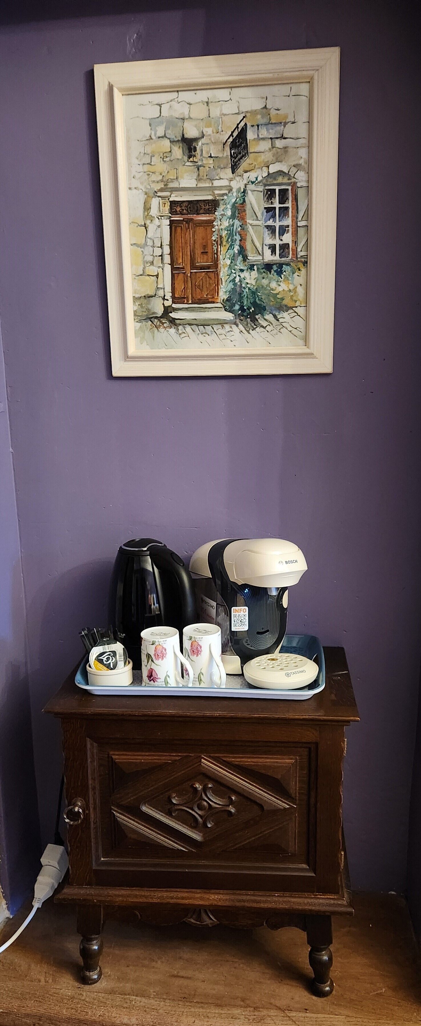 A framed painting of a rustic building, a coffee maker, a kettle, and a tray with cups and a teapot on a wooden cabinet against a purple wall.