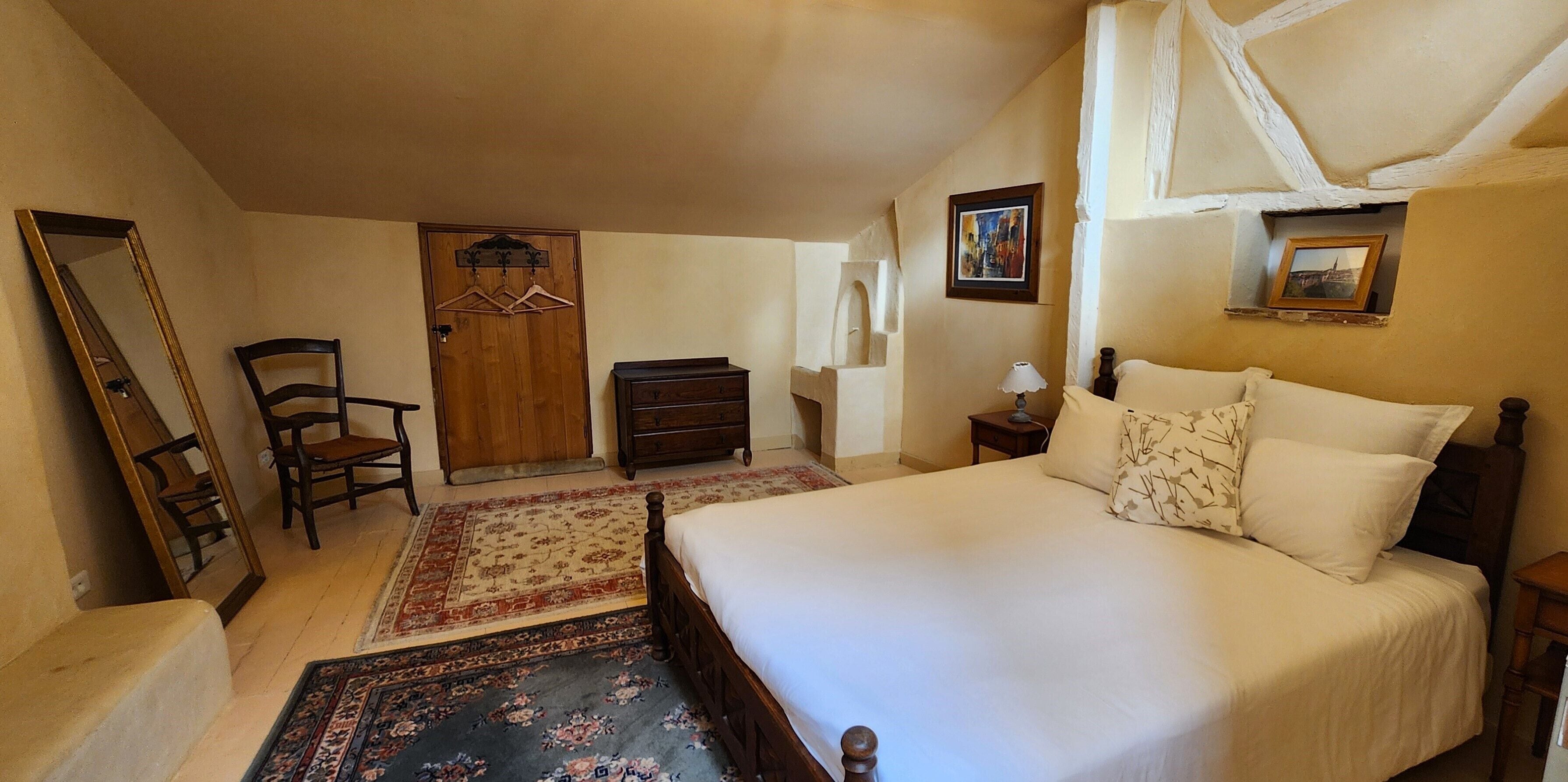 A bedroom with a large bed, a wooden dresser, a chair, a mirror, and a door with a coat rack.