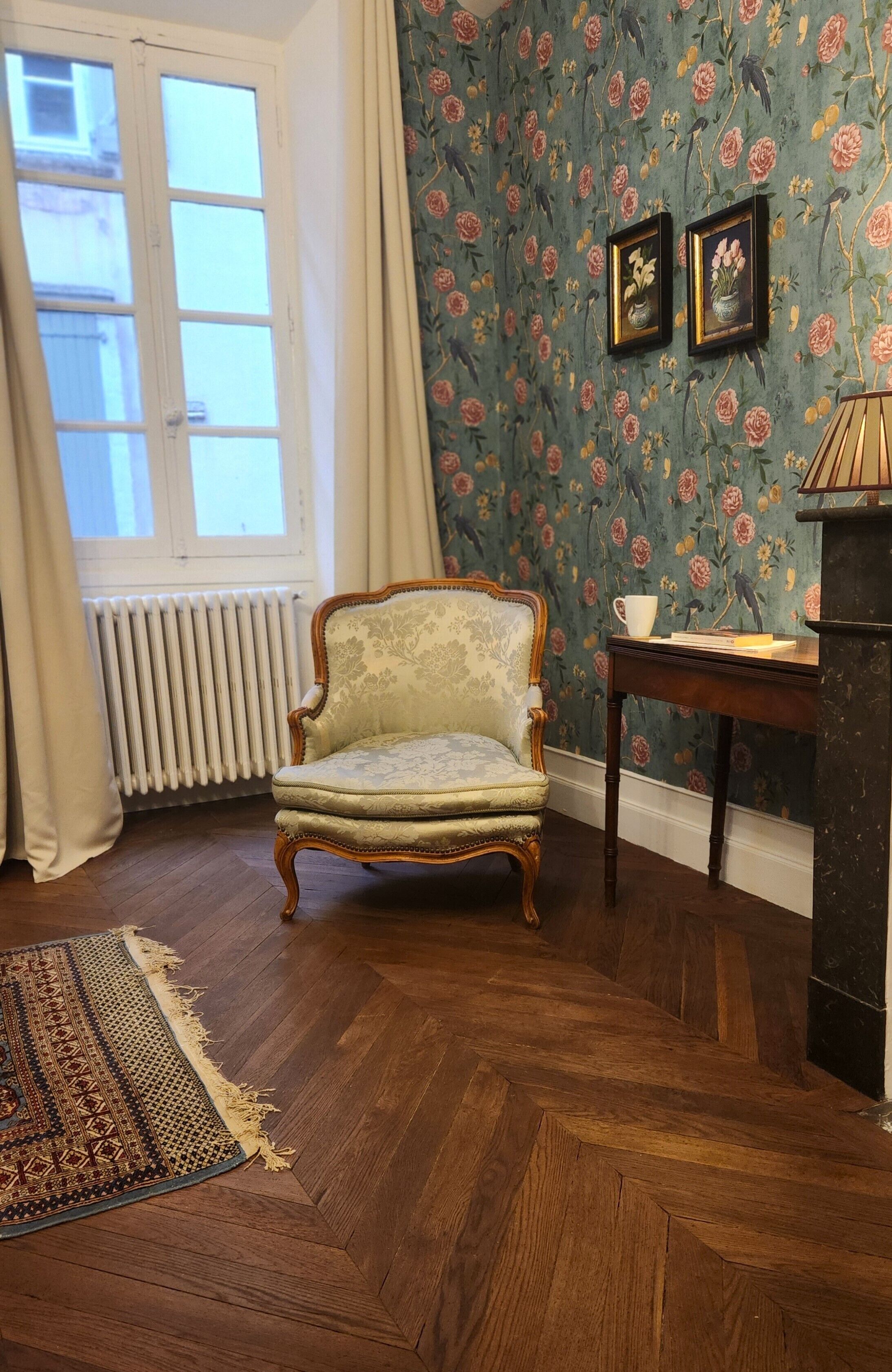 A room with a wooden herringbone floor, a vintage armchair, a radiator, a window with curtains, floral wallpaper, and a small table with a lamp.