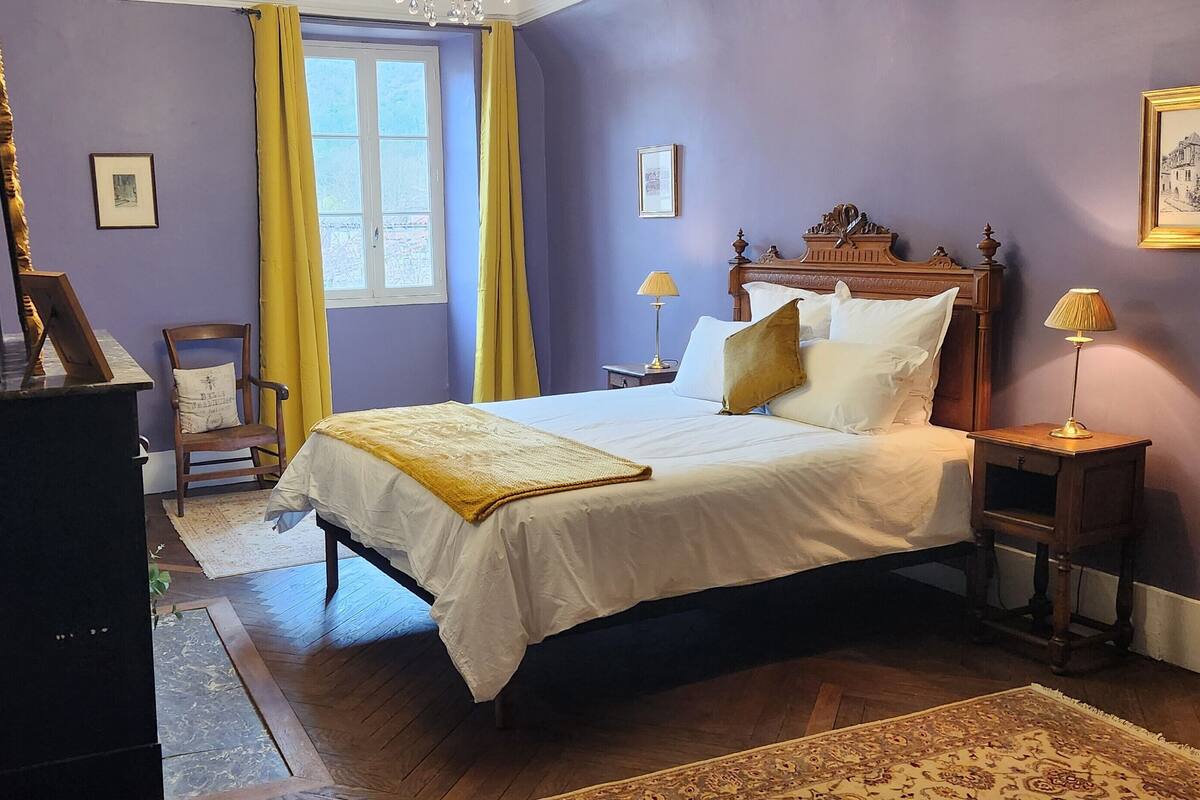 Une chambre à coucher avec un lit, une tête de lit en bois, deux tables de chevet avec des lampes, une fenêtre avec des rideaux jaunes et un tableau encadré au mur.