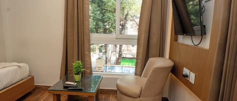 A room with a bed, a round chair, a small table with a plant, a TV mounted on the wall, and a window with curtains.