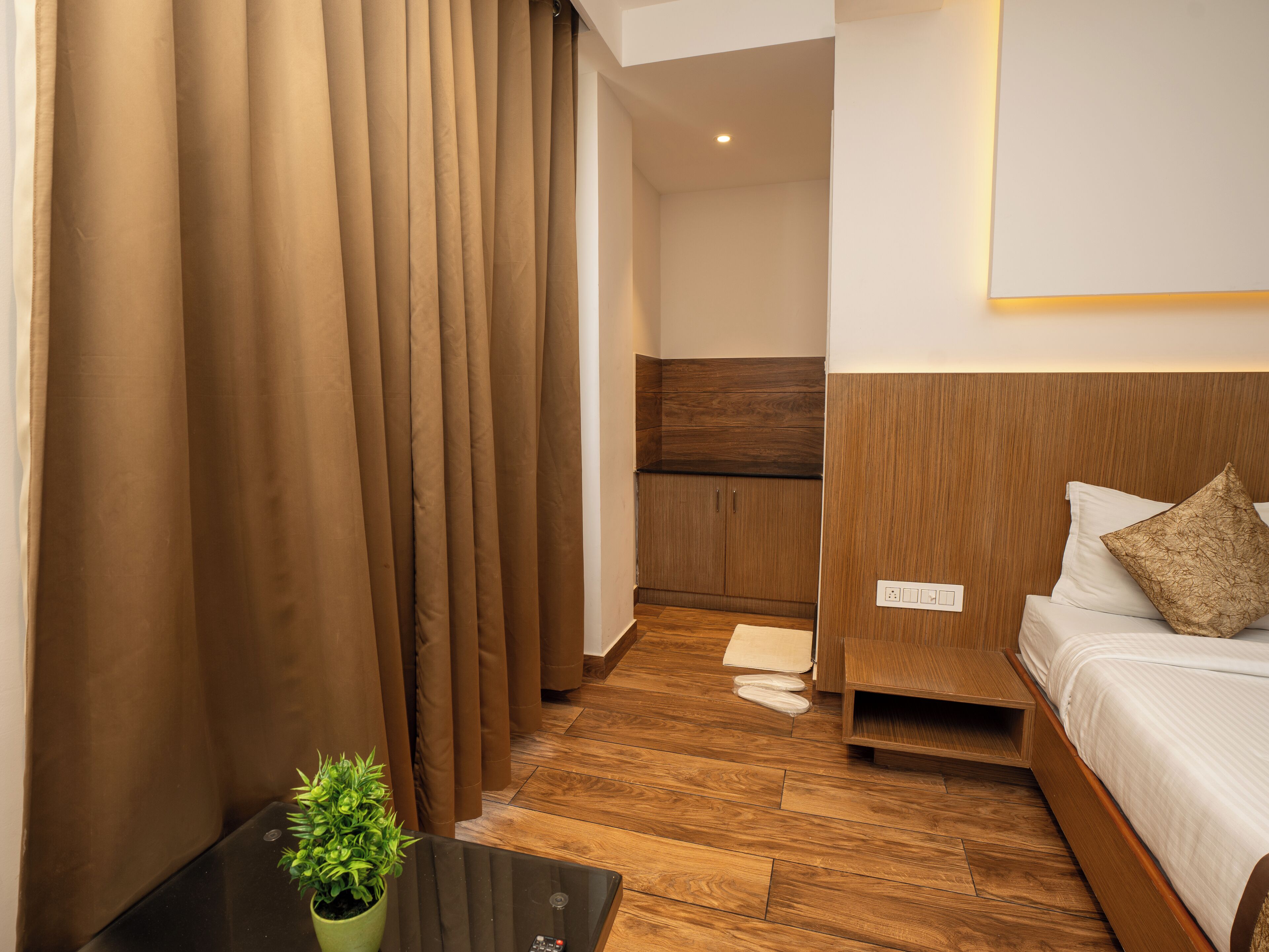 A hotel room with a wooden floor, a bed with white bedding, a small bedside table with a plant, and a curtain separating the room from the hallway.