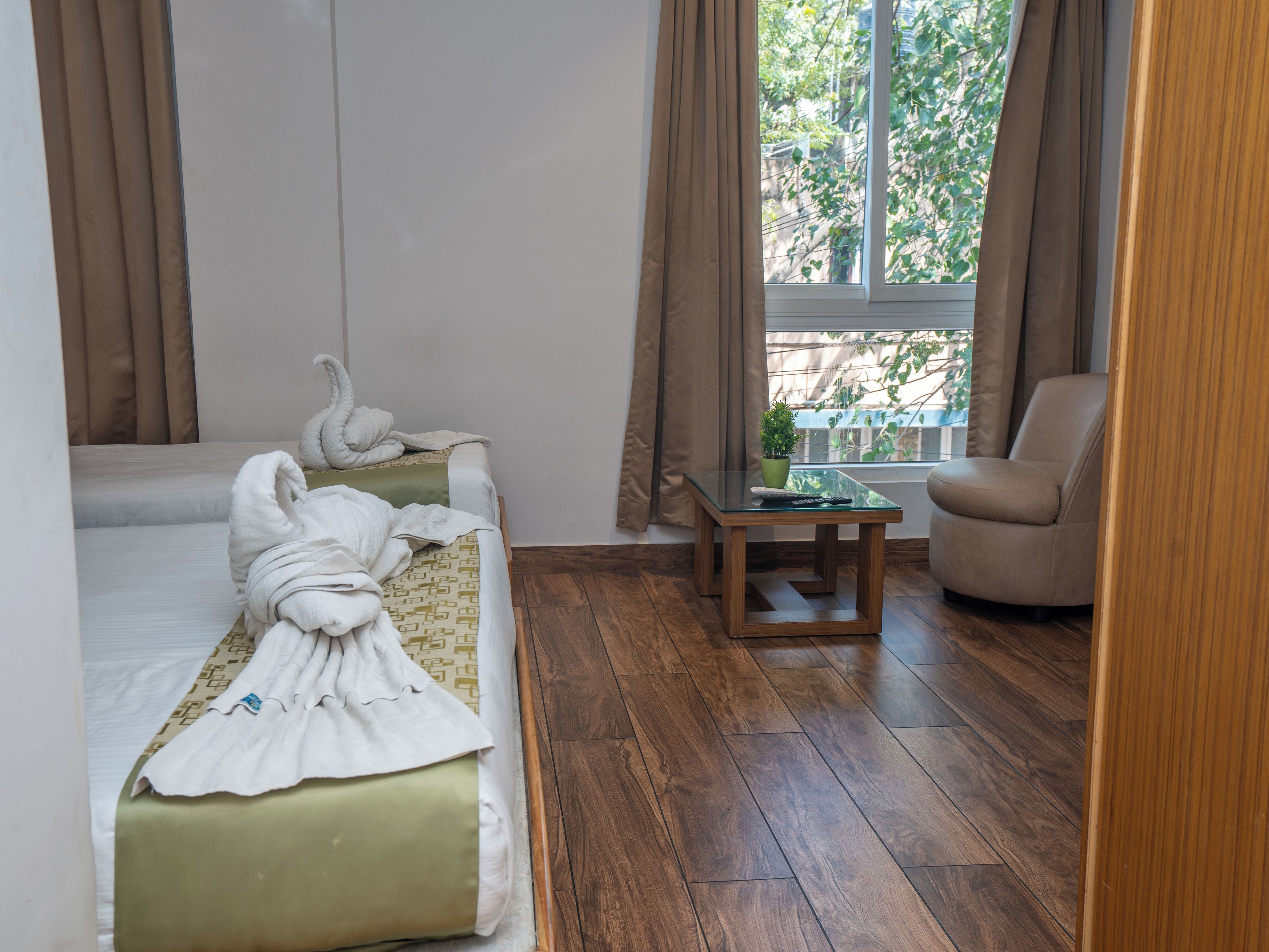 A hotel room with two beds, a wooden floor, a small table with a plant, and a view of trees through the window.