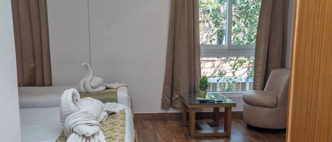 A hotel room with two beds, a wooden floor, a small table with a plant, and a view of trees through the window.