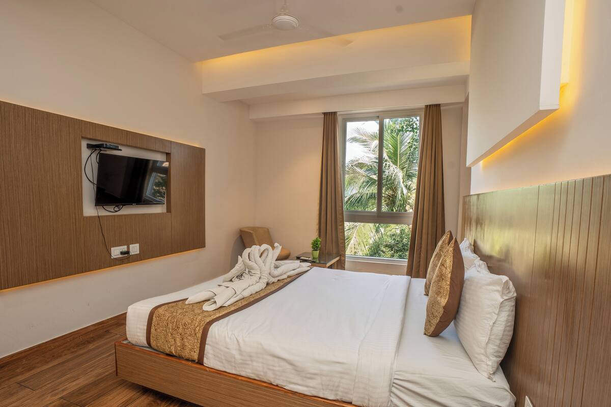 Une chambre d’hôtel moderne avec un lit en bois, un téléviseur à écran plat fixé au mur, une fenêtre avec des rideaux et une petite plante en pot.