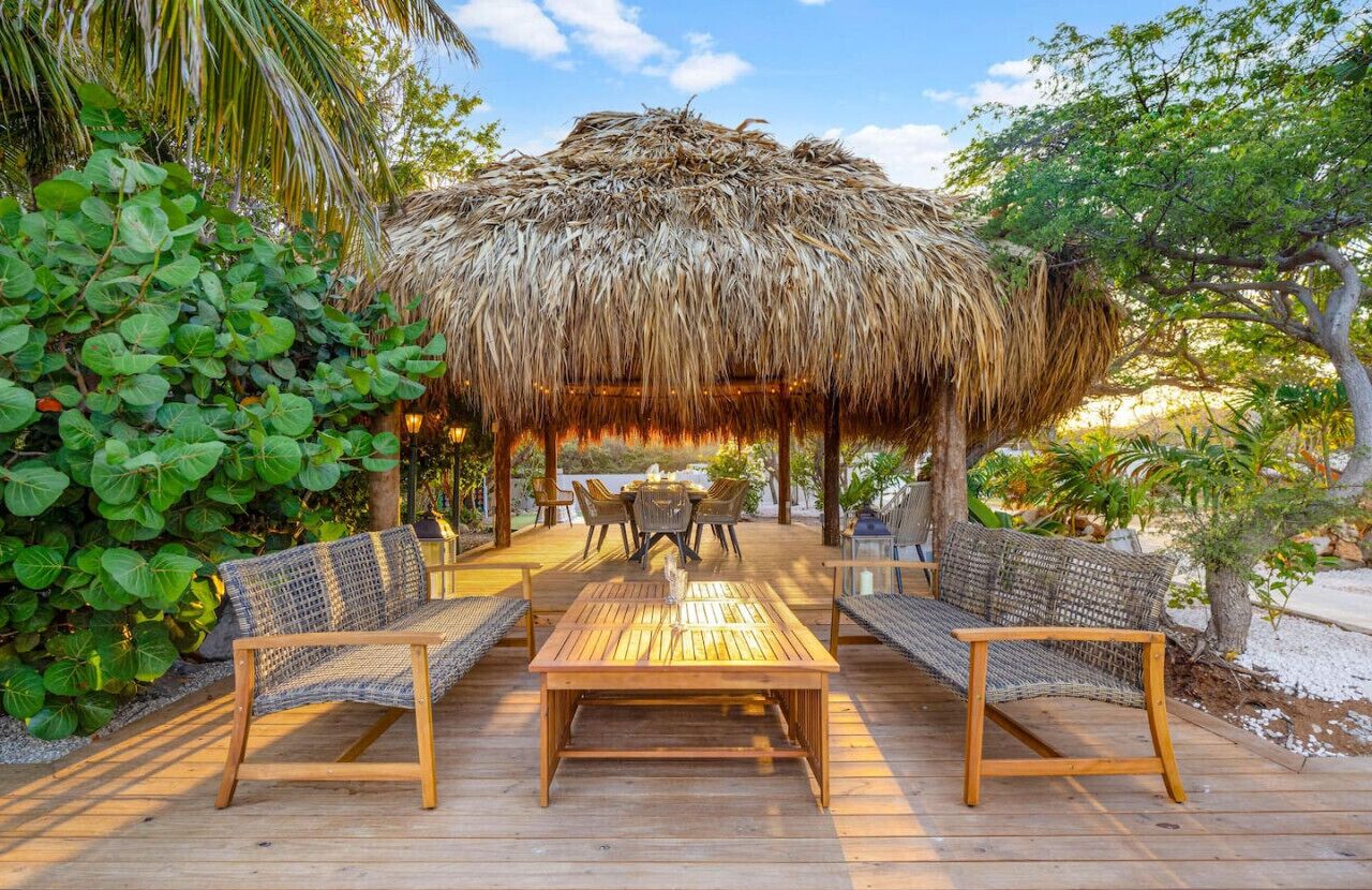 A wooden deck with wicker furniture, a thatched-roof structure, and lush greenery.