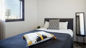 A neatly made bed with a patterned pillow, a bedside table, and a window with a view of buildings. - HaCarmel Market Vibrat Apt by Sea N Rent (Tel Aviv)