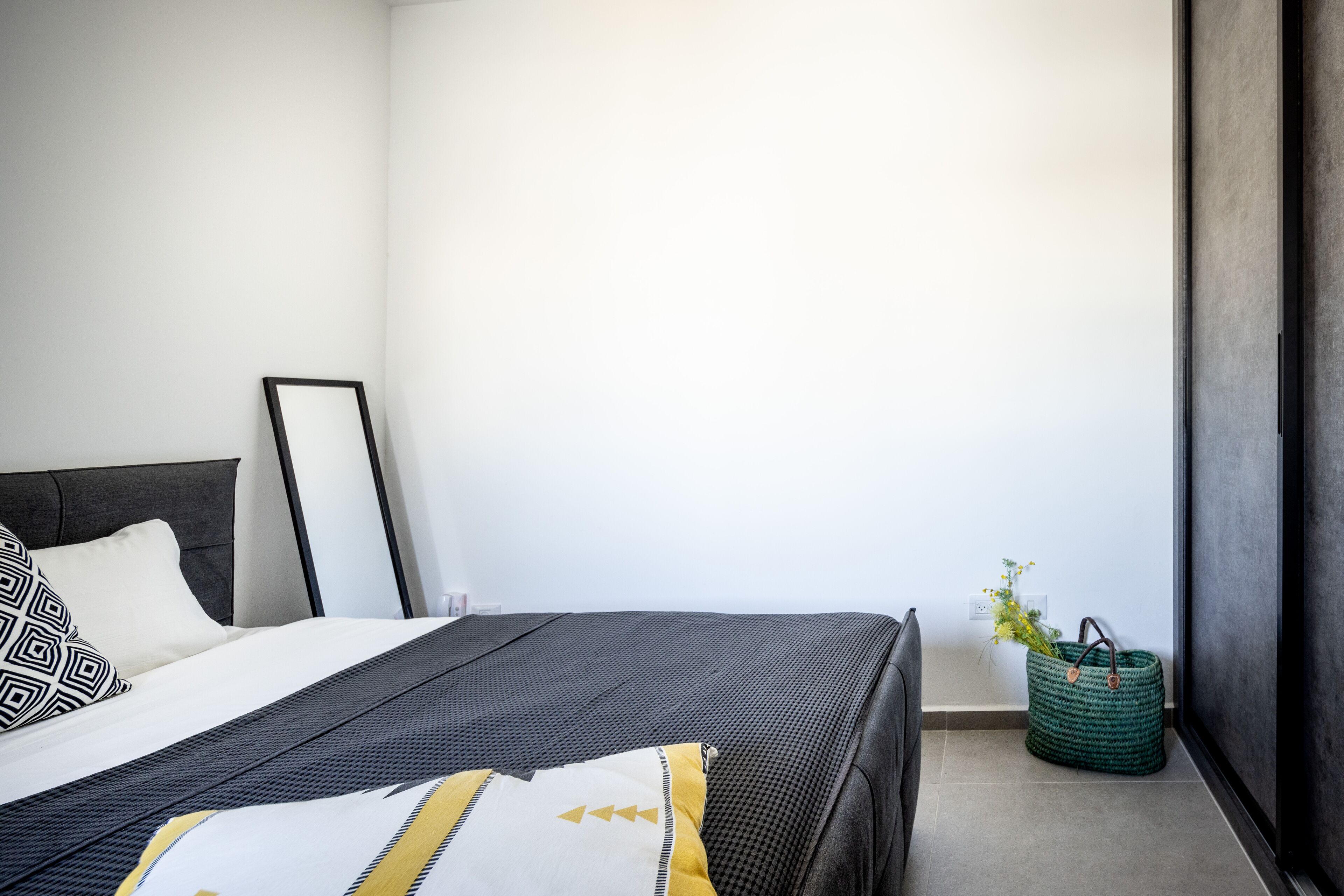 Une chambre à coucher moderne avec un lit, une table de chevet, un panier en osier vert et un luminaire fixé au mur.