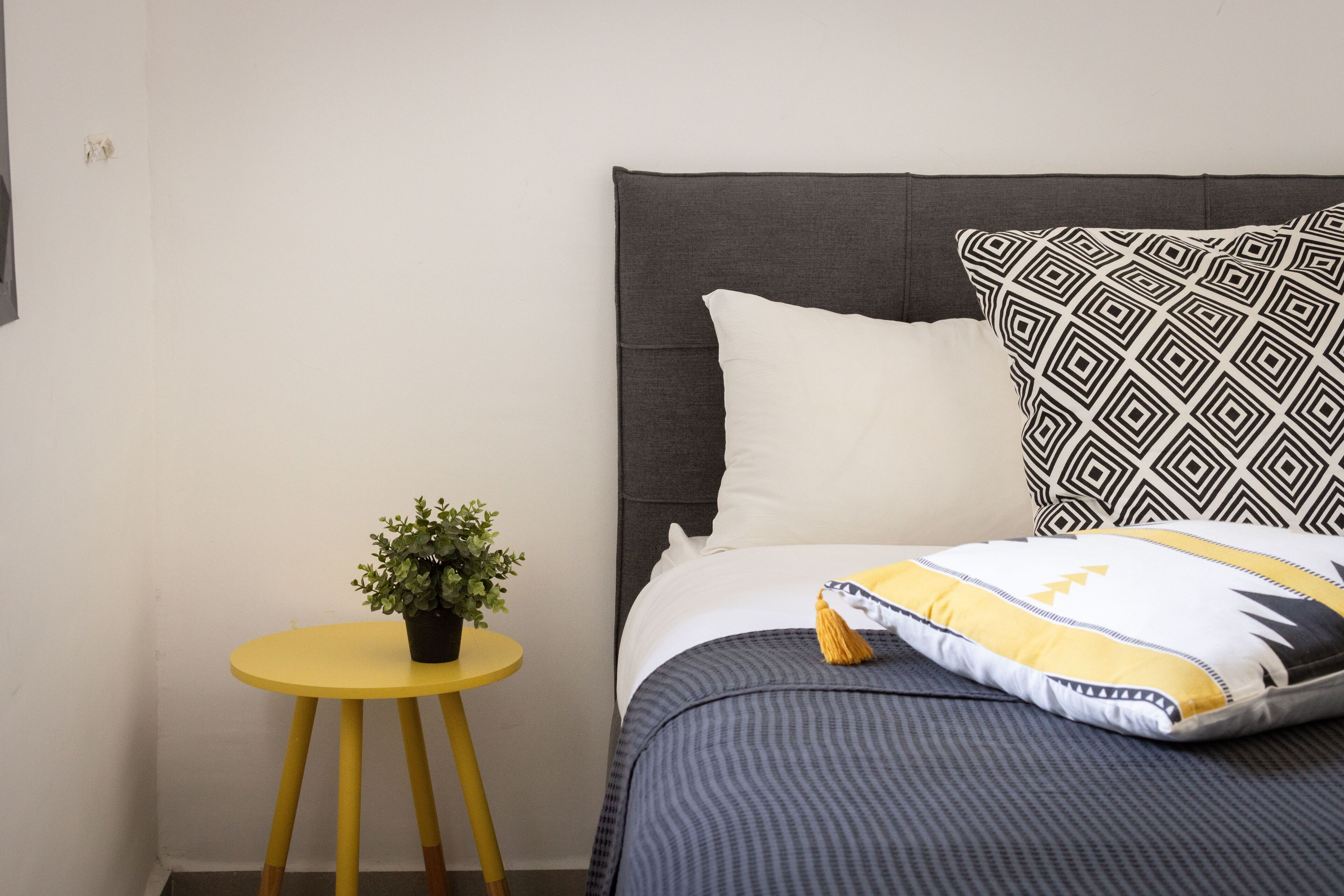 A neatly made bed with a gray headboard, a white pillow, a patterned pillow, and a striped blanket. There is a small round yellow table with a potted plant beside the bed.