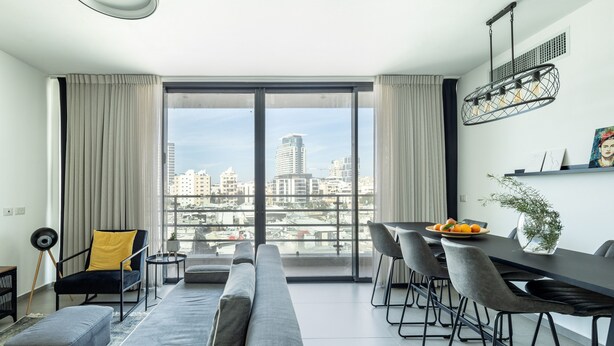 A modern living room with a large window, a sofa, a dining table, and a city view. - HaCarmel Market Vibrat Apt by Sea N Rent (Tel Aviv)