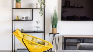 A modern living room with a yellow wicker chair, a grey sofa, a black metal shelving unit, and a potted plant. - HaCarmel Market Vibrat Apt by Sea N Rent (Tel Aviv)