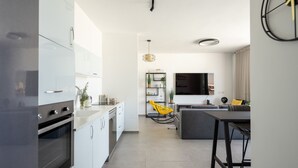 A modern kitchen with white cabinets, a built-in oven, and a dining area with a grey sofa and a yellow chair. - HaCarmel Market Vibrat Apt by Sea N Rent (Tel Aviv)