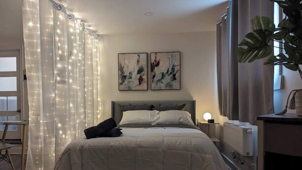 A bedroom with a bed, curtains, a bedside table with a lamp, and two abstract paintings on the wall. - Stylish Studio, Steps to Transit (Montréal)