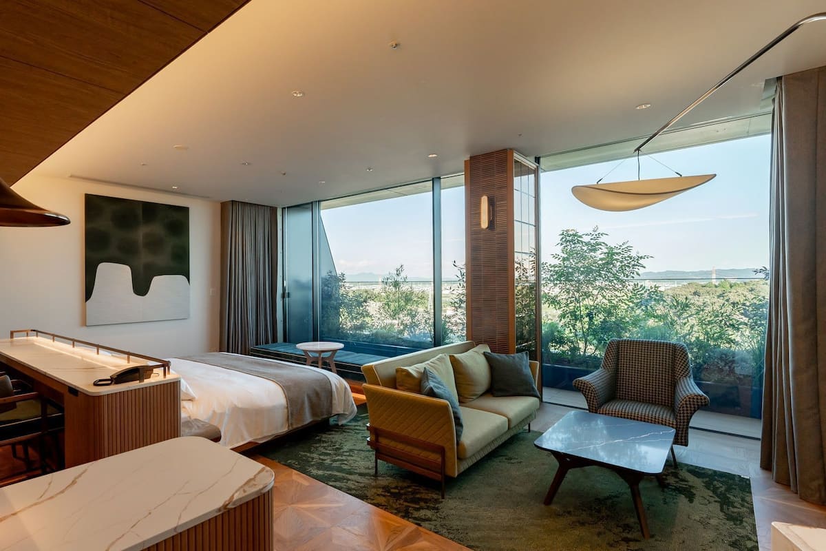 A modern hotel room with a bed, sofa, armchair, and a dining area with a table and chairs.