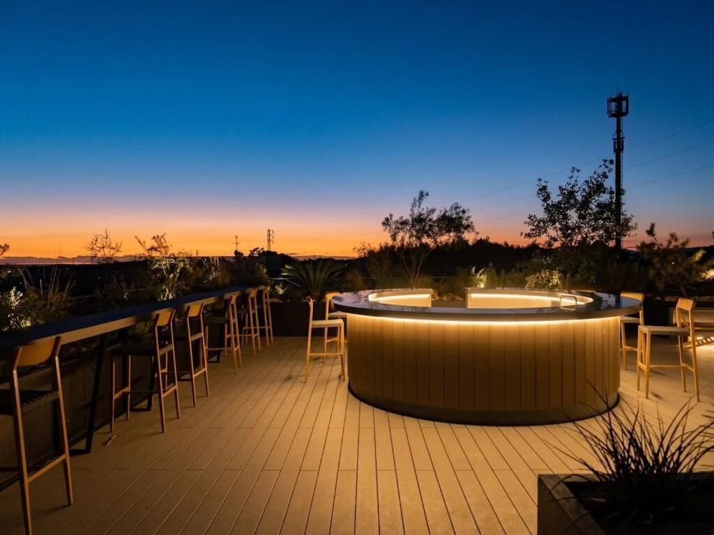 Un espace bar circulaire avec une table de billard et des tabourets de bar, entouré d’une terrasse en bois et de plantes.