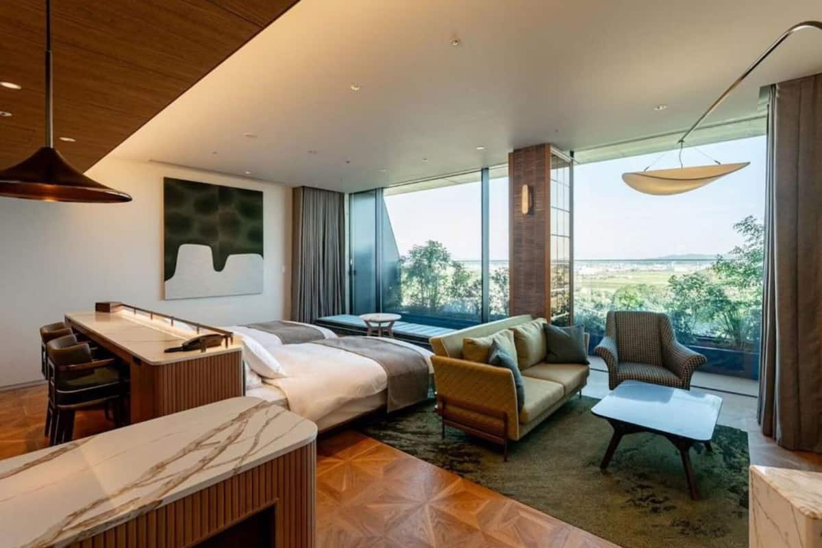 A modern hotel room with a large bed, a desk, a sofa, and a view of the outdoors.
