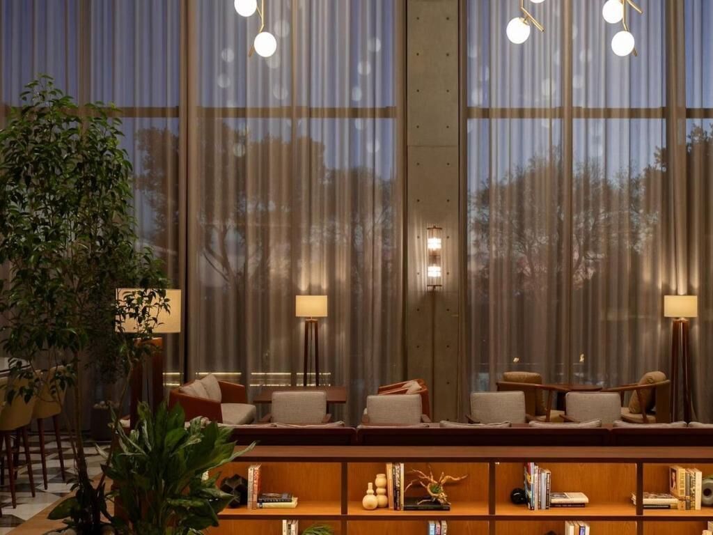 A modern hotel lobby with a seating area, large windows, and a bookshelf.