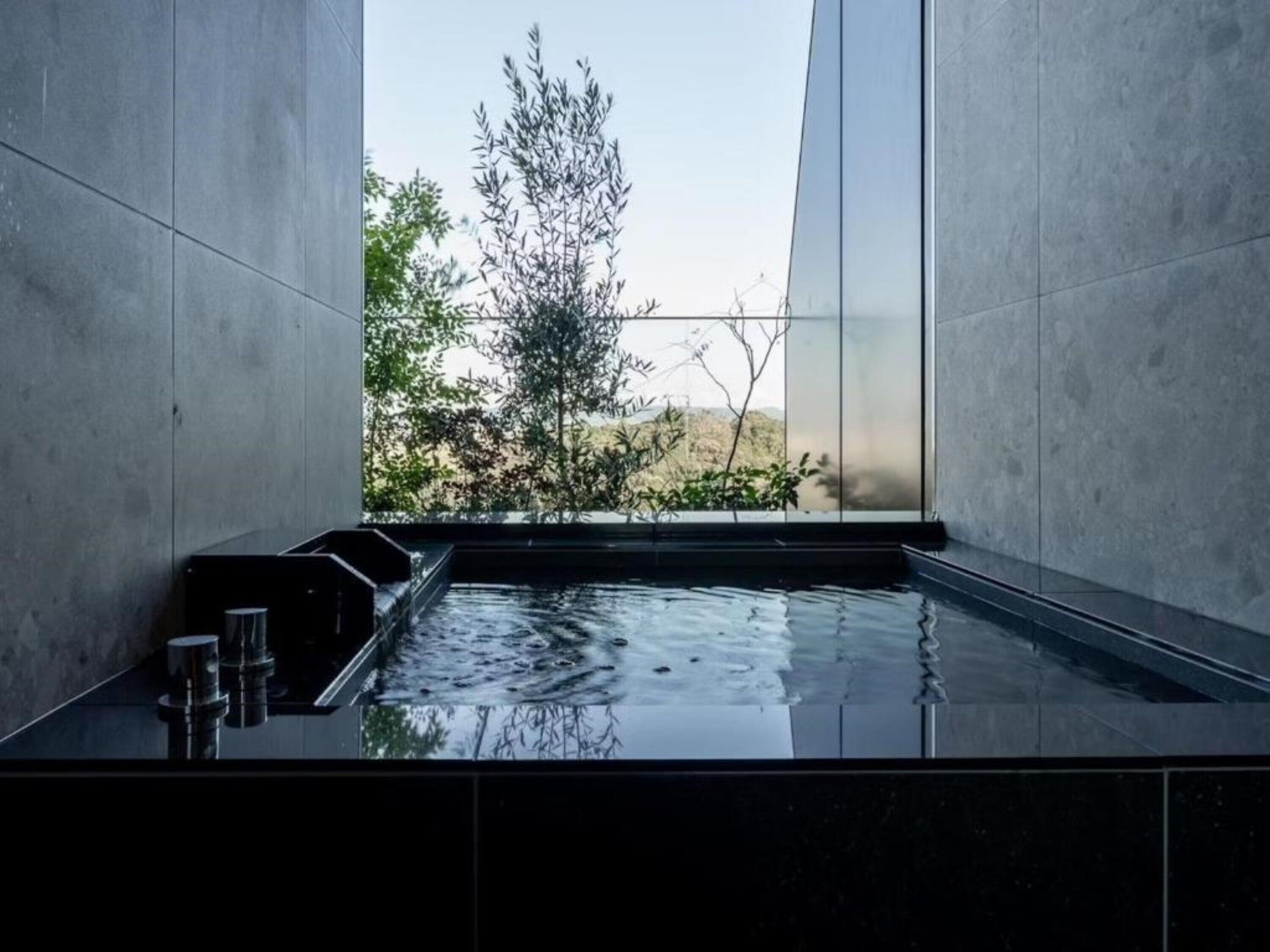 Un spa moderno con una bañera de hidromasaje cubierta de vidrio, rodeada de muros de concreto y vistas a árboles y colinas.