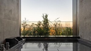 Un baño moderno con un espejo grande, una encimera de mármol negro y vistas a un jardín con hierbas altas y árboles.