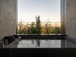 Um banheiro moderno com um espelho grande, bancada de mármore preto e vista para um jardim com gramíneas altas e árvores.