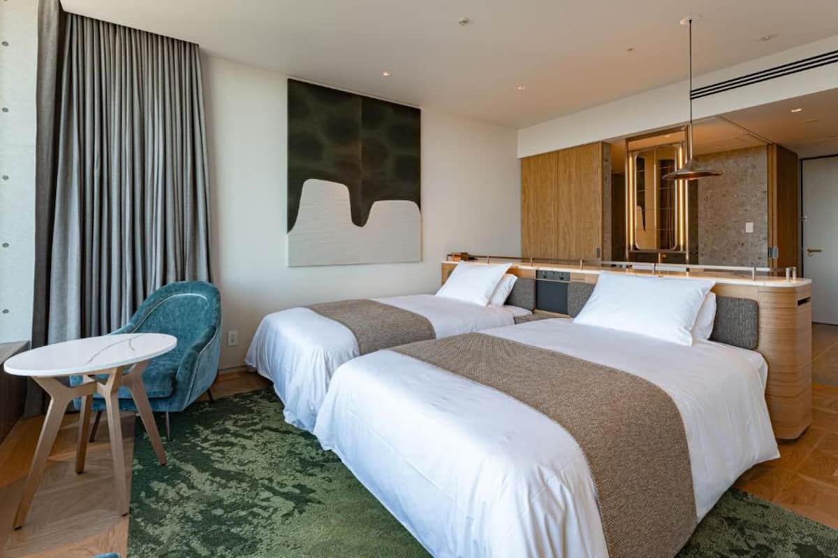 A hotel room with two beds, a small table, a chair, a large abstract painting, and a modern bathroom with a mirror and shower.