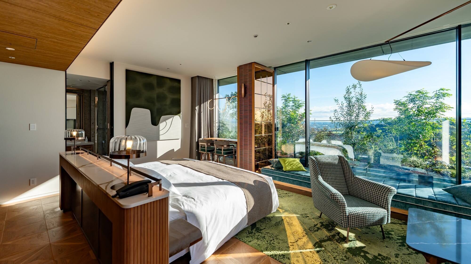 Een moderne slaapkamer met een groot bed, een bureau, een stoel en uitzicht op groen door grote ramen.