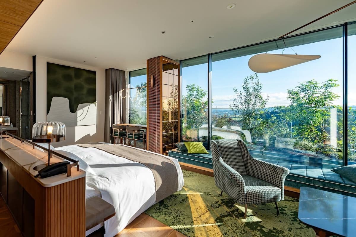 A modern bedroom with a large bed, a desk, a chair, and a view of greenery through large windows.