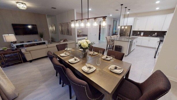 A modern dining area with a long table set for six, a sofa, and a kitchen in the background. - Luxury 6B Pool Home At Storey Lake (Kissimmee)