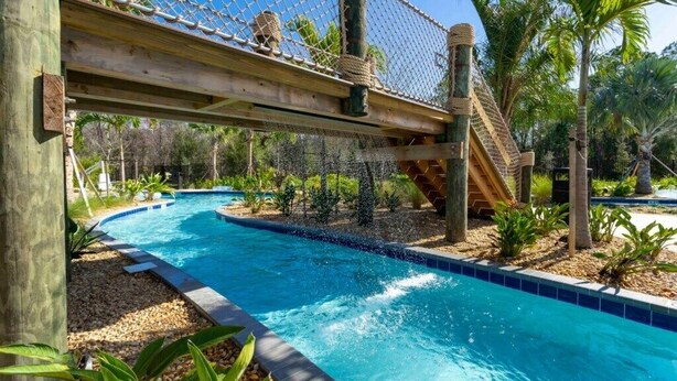 Una piscina con un puente colgante y un tobogán, rodeada de palmeras y plantas.