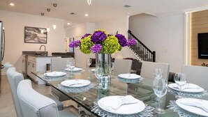 Un moderno comedor con una mesa de vidrio dispuesta para seis personas, rodeado de sillas blancas. Un jarrón con flores moradas y amarillas está sobre la mesa.