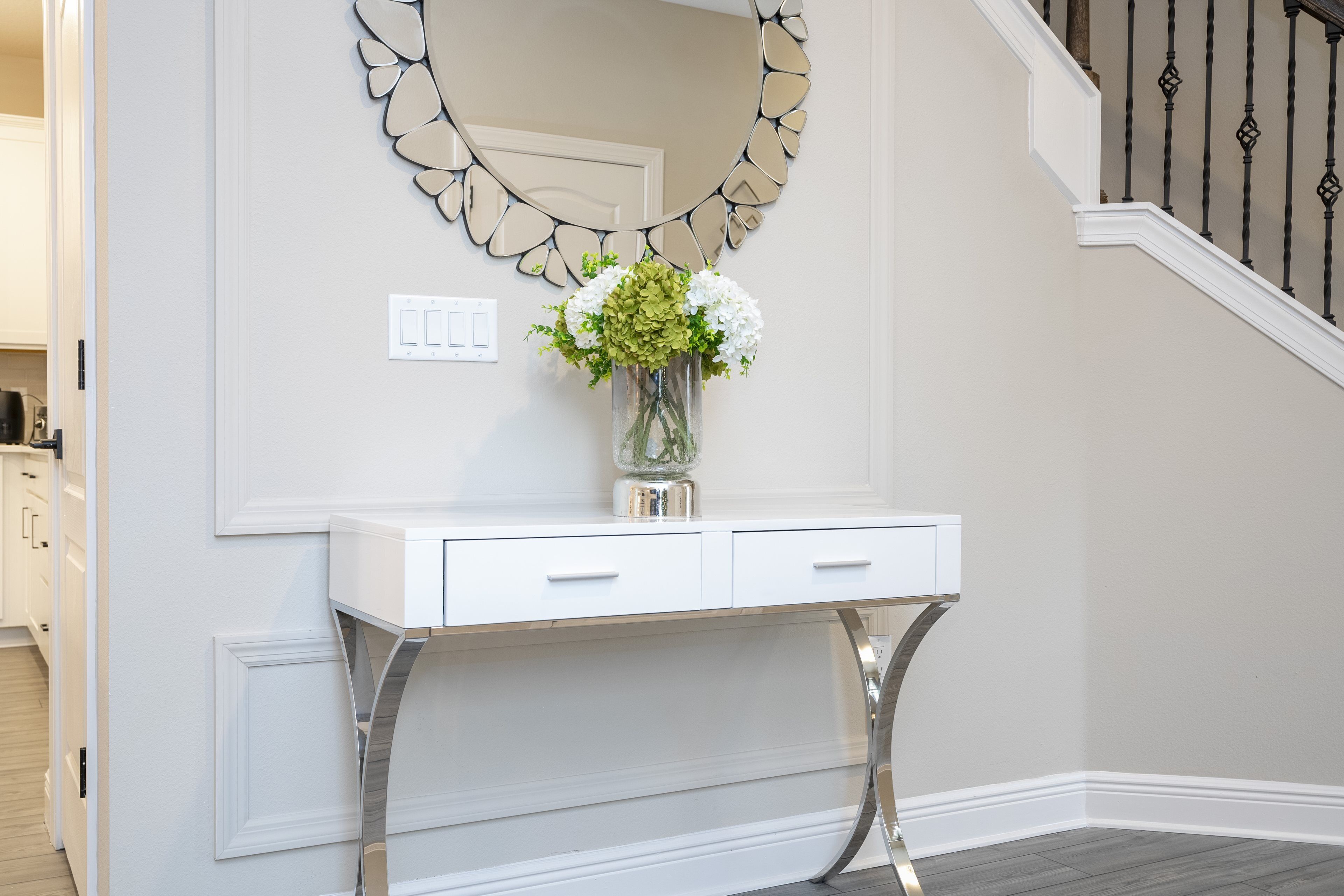 Une console blanche, ornée d’un vase de fleurs, d’un miroir décoratif et d’un interrupteur sur le mur.