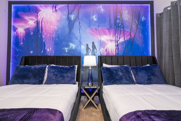 Una cama doble con colcha blanca y morada, dos almohadas azules mullidas, una mesita de noche con lámpara y un mural en la pared con una escena de bosque.