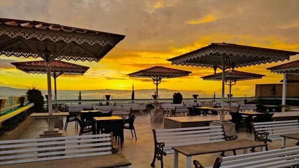Uma área externa com mesas e bancos, guarda-sóis e vista para o pôr do sol sobre a água.