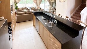 Una cocina moderna con encimera negra, armarios de madera y un lavamanos integrado. Hay una zona de comedor con una mesa y sillas, y una escalera que conduce a otro nivel.
