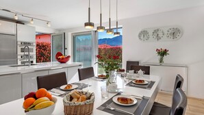 A modern kitchen with a large island, a dining area with a table set for two, and a view of a mountain and flowers through the windows. - Villa Dolce Vita With Private Pool (Lugano)