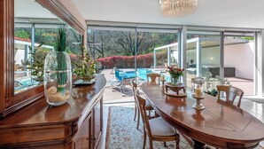 A dining area with a wooden table, chairs, and a chandelier. There is a view of a swimming pool and outdoor seating. - Villa Dolce Vita With Private Pool (Lugano)