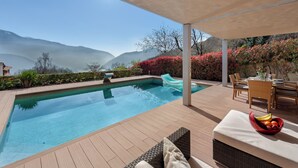 A pool area with a view of mountains, a dining table with chairs, and a fruit bowl on a cushioned bench. - Villa Dolce Vita With Private Pool (Lugano)
