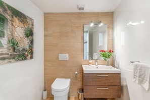 A modern bathroom with a beige tiled wall, a white toilet, a wooden vanity with a mirror, and a vase with red flowers.