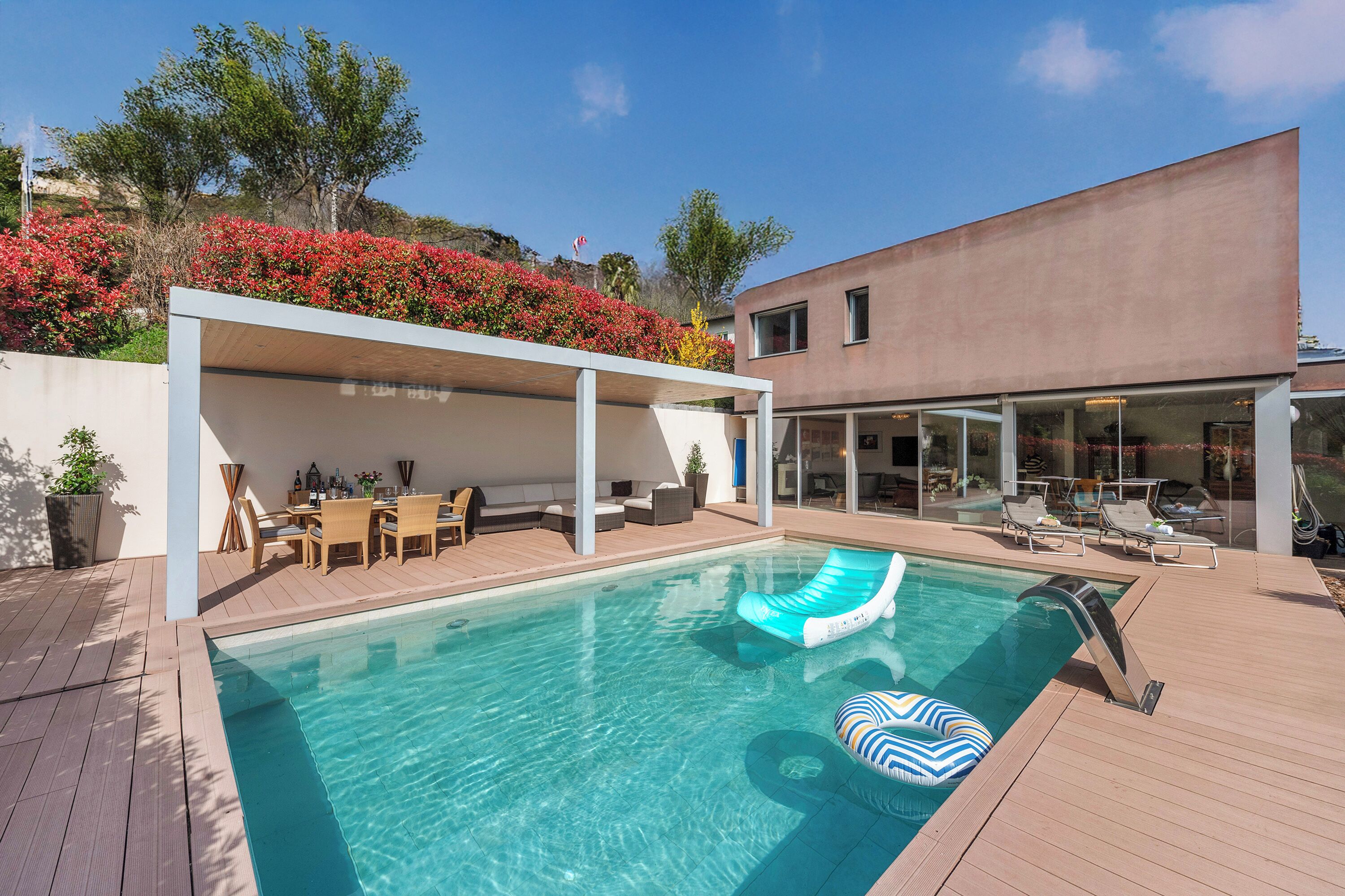 Ein moderner Poolbereich im Freien mit Holzdeck, Liegestühlen und einer überdachten Sitzecke.