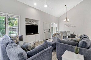 A modern living room with grey sofas, a flat-screen TV, white cabinetry, and a dining area with a chandelier. - Walk to Beach & Golf | 2 BR Palmetto Dunes Villa (Hilton Head Island)