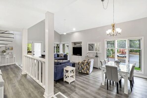 A modern interior with a dining area, kitchen, and living space, featuring a chandelier, large windows, and a staircase.