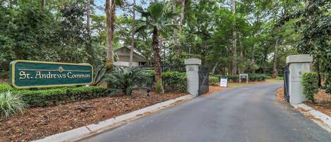 A gated entrance to St. Andrews Commons with a sign, a paved driveway, and surrounding greenery.