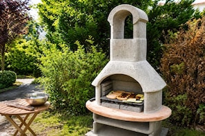Un barbecue extérieur en pierre, doté d’une cheminée, entouré de verdure.
